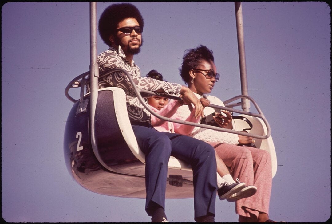 A man, woman, and young girl sit together on a ski lift or amusement park ride, wearing sunglasses and casual 1970s clothing, against a clear blue sky. The girl sits between the adults, partially hidden.