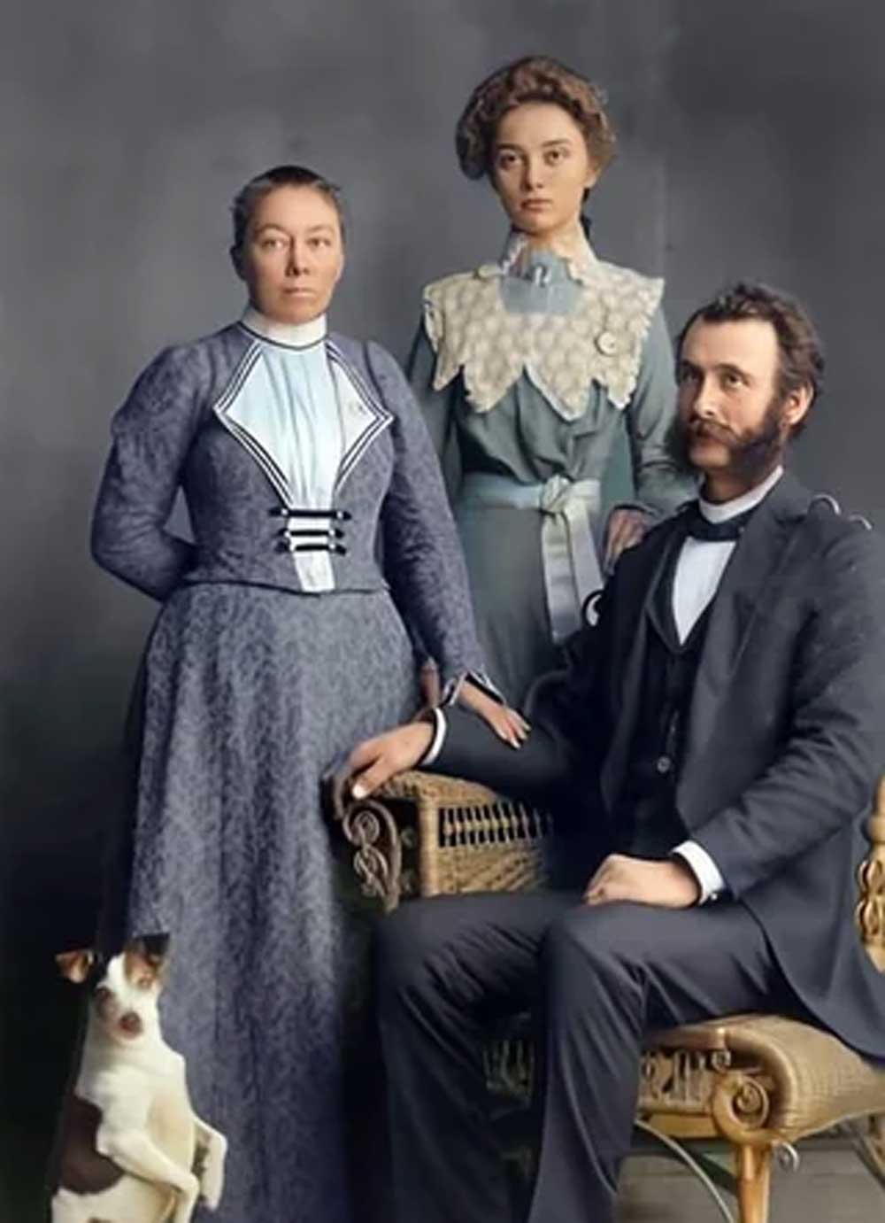 A vintage family portrait shows a woman and a young woman standing behind a seated man in a suit, with a small dog sitting upright at their feet. All are dressed in late 19th or early 20th-century clothing.