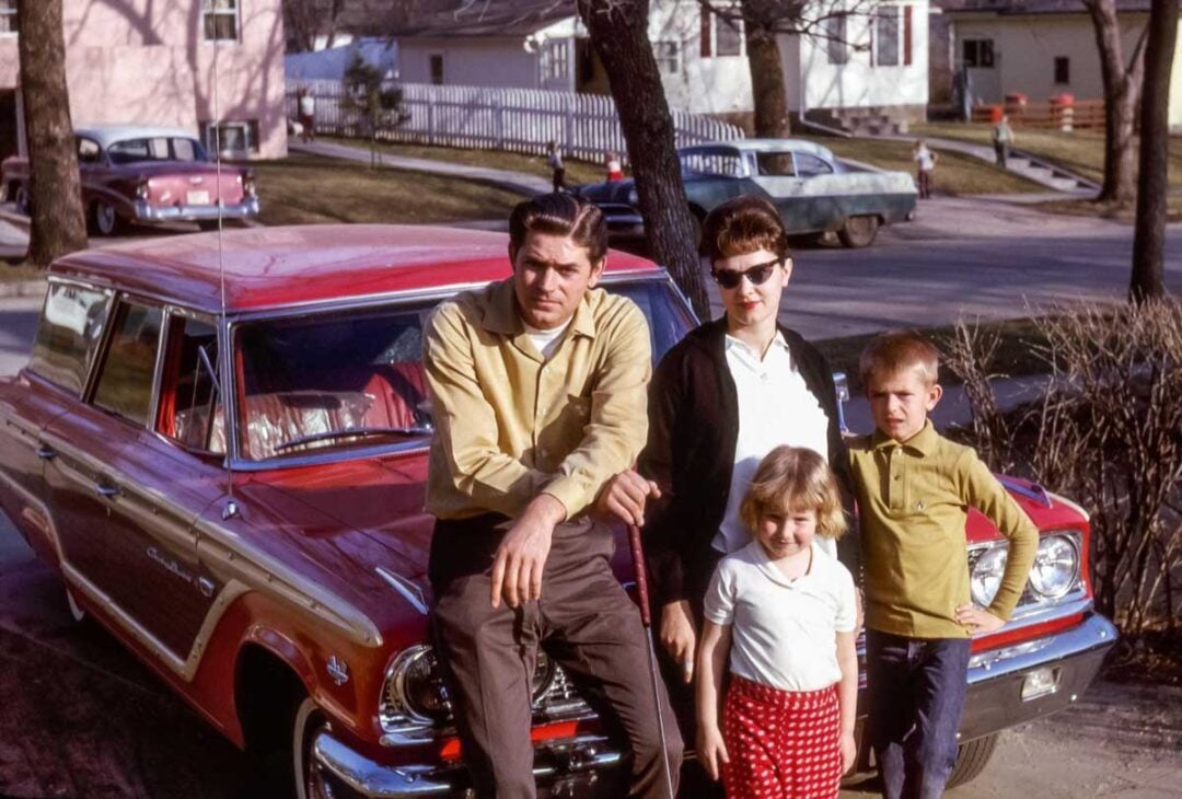 A man, woman, young girl, and young boy pose in front of a red vintage car parked on a suburban street lined with houses and trees on a sunny day.