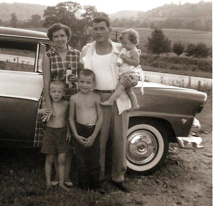 A black-and-white photo of a family of five standing in front of a classic car; a man holds a young child, with a woman and two boys beside them, in a rural outdoor setting with trees and hills in the background.