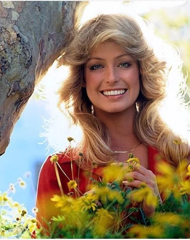 A woman with blonde, feathered hair and a bright smile stands outdoors near a tree, surrounded by yellow flowers and greenery, with sunlight shining behind her.