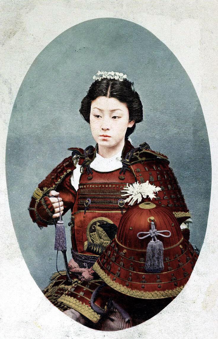 A Japanese woman wearing traditional samurai armor with floral hair ornaments, posed against a pale background. An ornate red helmet rests in front of her. The image has a vintage, hand-colored appearance.