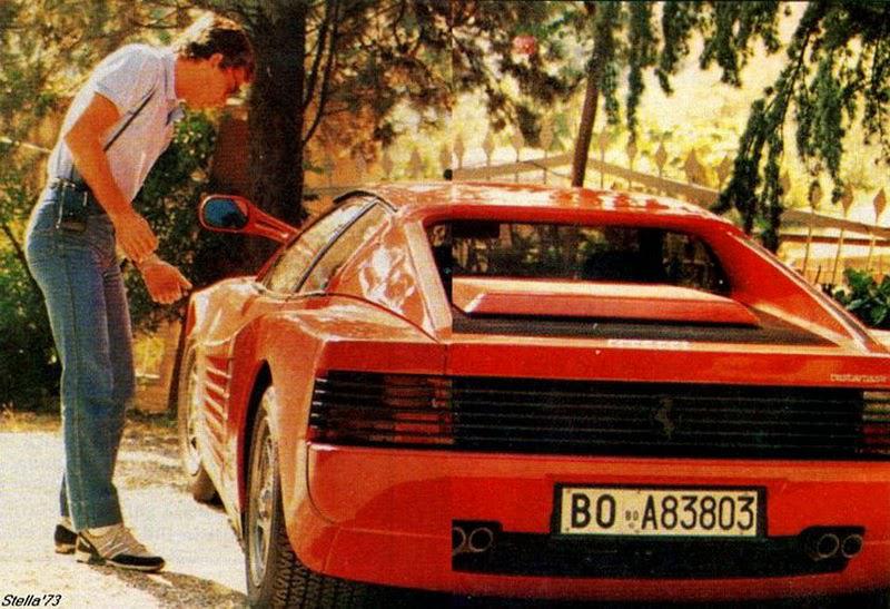 A person in jeans and a white shirt stands beside a red Ferrari Testarossa, looking into the open driver's door. The car is parked outdoors under trees, with sunlight creating a dappled effect.
