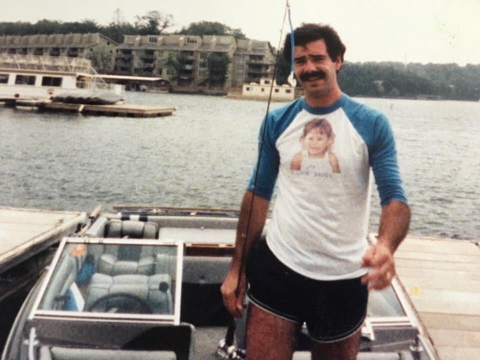 A man with a mustache stands on a docked boat, holding a fishing rod. He wears a blue and white shirt featuring a child’s photo and black shorts. In the background, there is a marina and lakeside buildings.