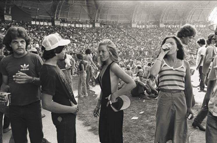 A black-and-white photo of young people at a crowded stadium event. Three stand in the foreground, one drinking from a can, while others sit and gather on the grass with the audience filling the stands behind them.