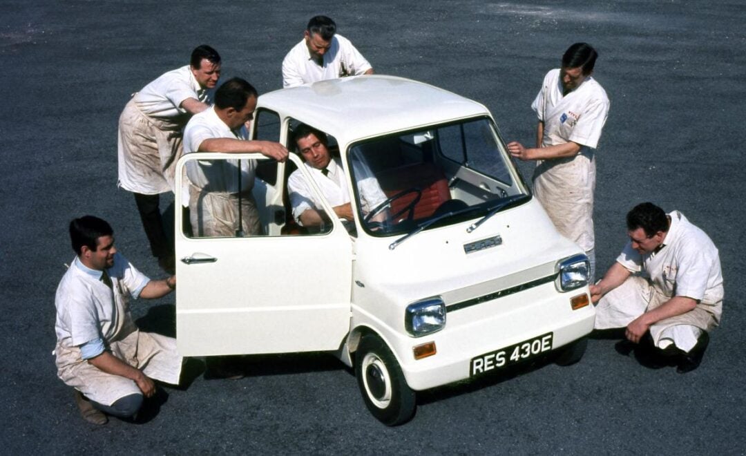 Six men in white shirts and aprons surround a small white vintage car with one man sitting inside. The car is parked on a paved surface, and the men appear to be examining or presenting it.