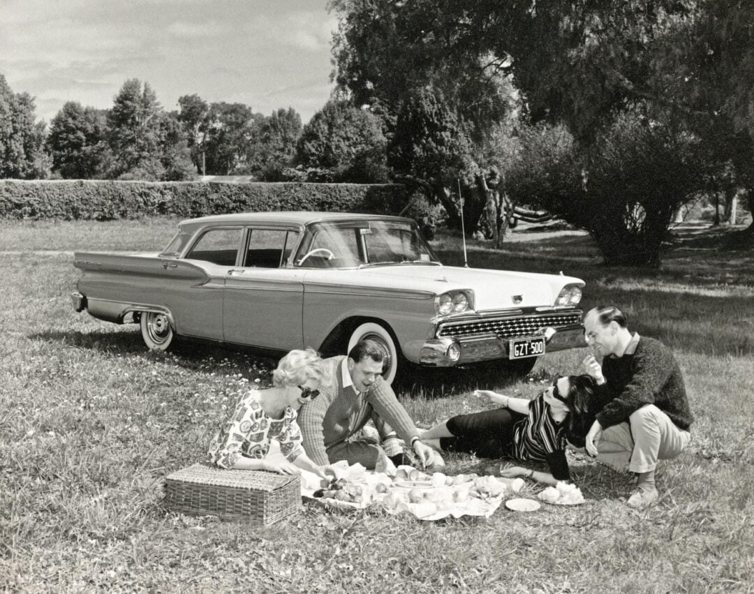 Four people have a picnic on a blanket in a grassy park, with food spread out beside a wicker basket. A vintage car is parked nearby under leafy trees, and the scene looks relaxed and cheerful.