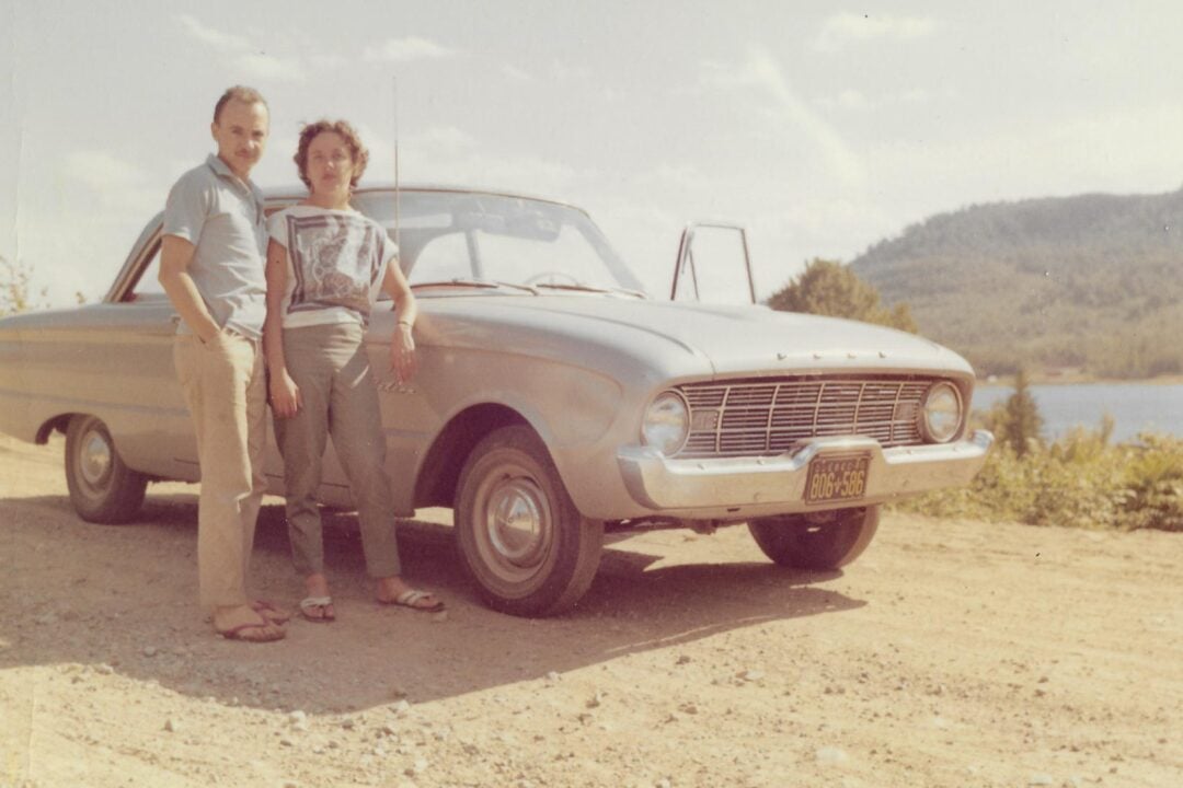 A man and woman stand beside a vintage silver car on a dirt road, with a lake and tree-covered hills in the background on a sunny day.