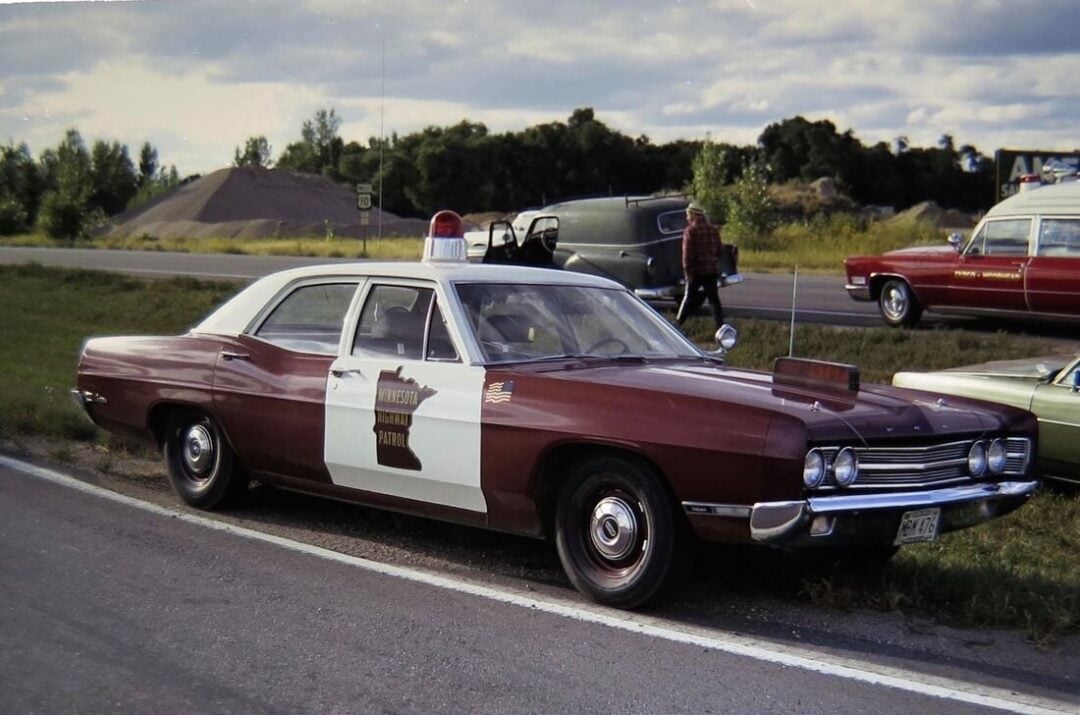 Vintage brown and white police car with a roof-mounted siren parked on the roadside, displaying a "Minnesota State Patrol" emblem on the door. Other classic vehicles and trees are visible in the background.