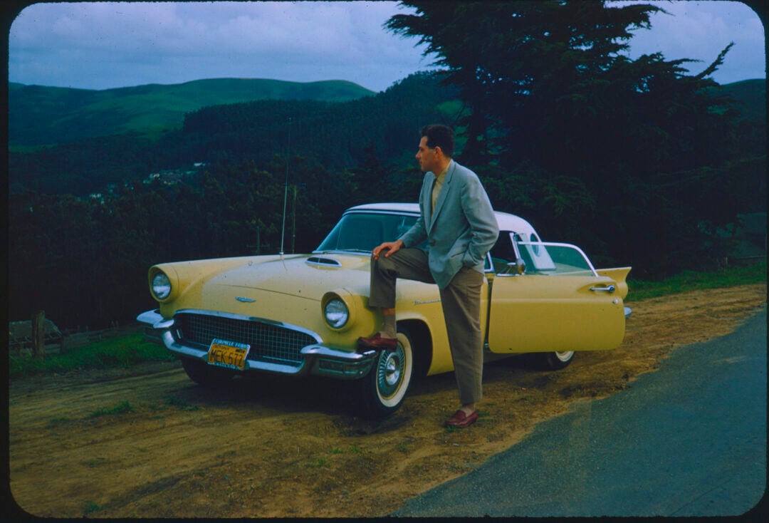 A man in a suit leans with one foot on the bumper of a yellow vintage car parked on a roadside, with green hills and trees in the background under a cloudy sky.
