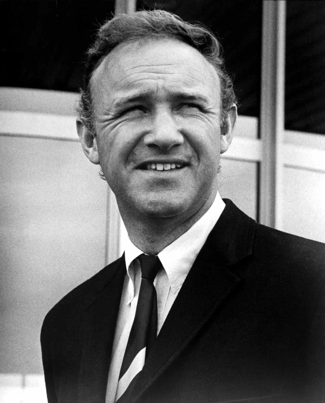 Black and white photo of a man in a suit and tie, standing outdoors and looking slightly to his left with a neutral expression. The background shows large windows or glass doors.