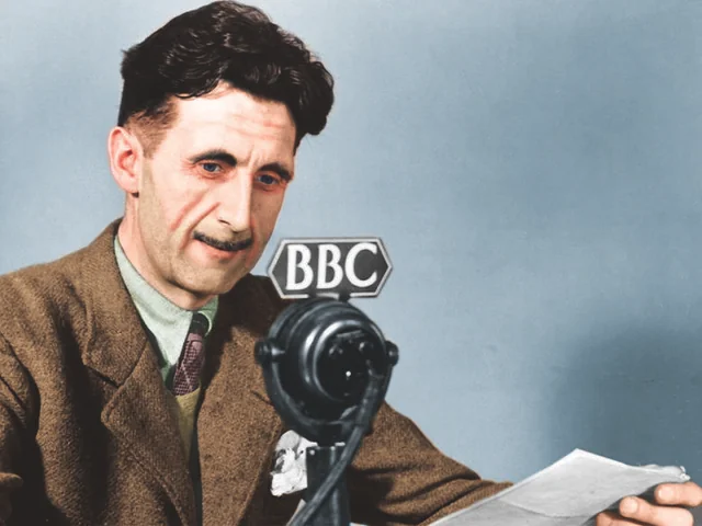 A man in a brown suit speaks into a vintage BBC microphone while holding papers, seated against a plain blue background.