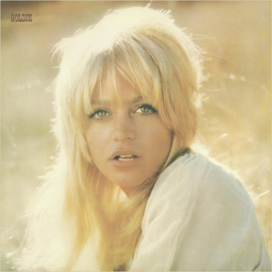 A blonde woman with long hair and bangs looks toward the camera in soft, golden light. She wears a light-colored top, and the background is blurred and sunlit. The word "GOLDIE" appears in the upper left corner.