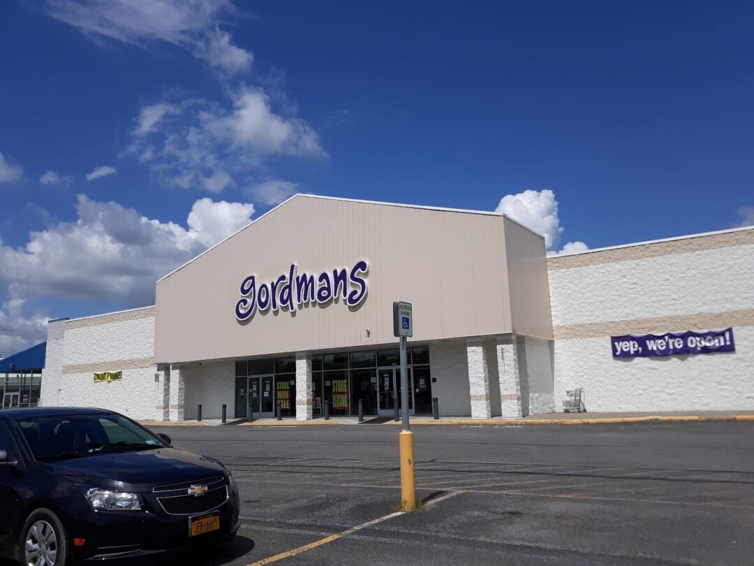 A Gordmans store with a "yep, we're open!" banner on the front. A black car is parked in the lot, and the sky is partly cloudy.