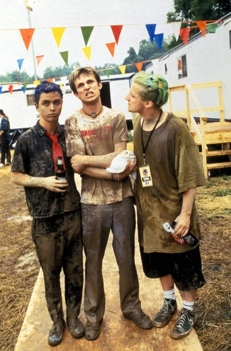 Three young men with colorful hair and muddy clothes stand outdoors at a festival or event, surrounded by colorful flags and trailers, looking tired but expressive. One person has his arms crossed and a tense expression.
