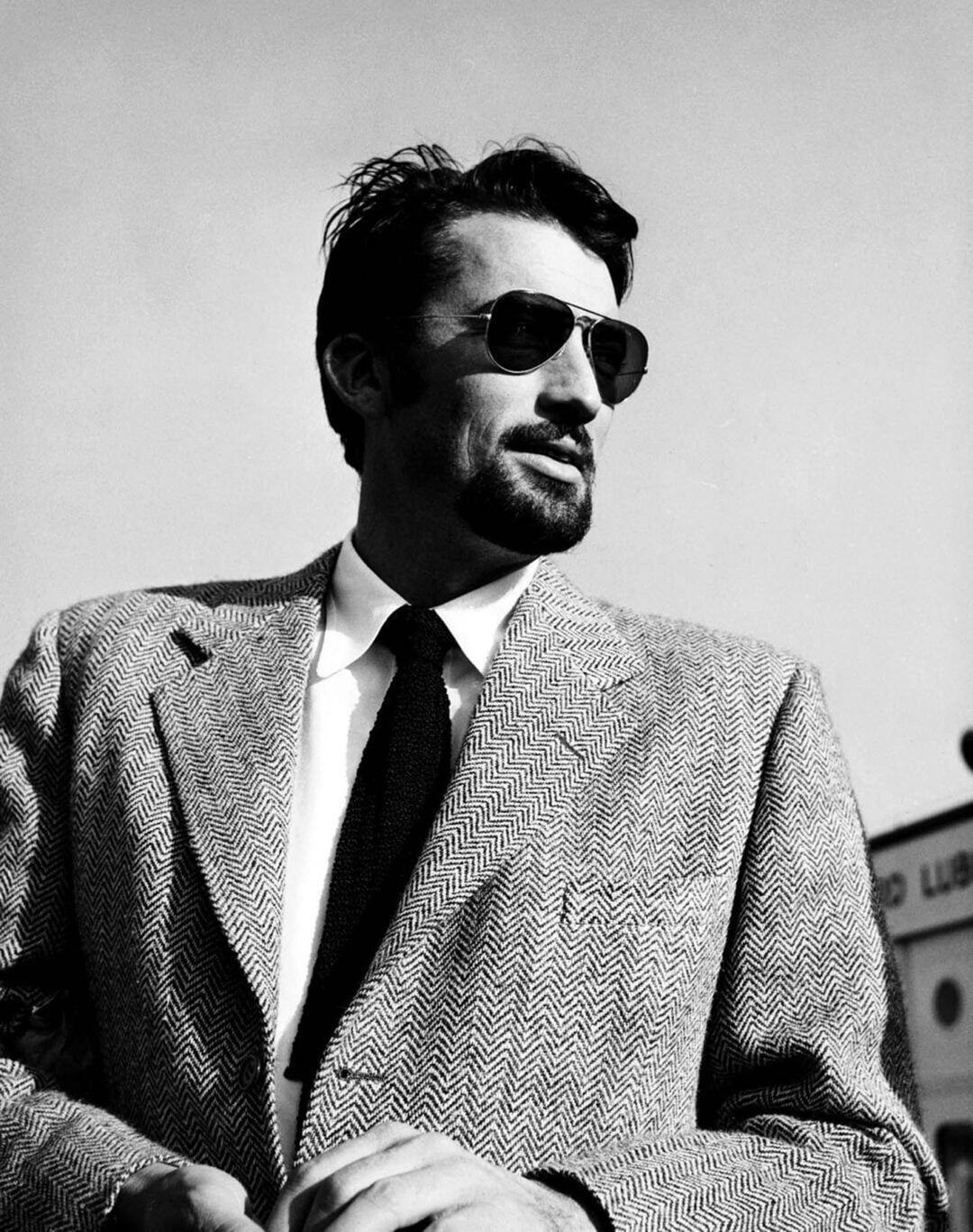 A man in a herringbone suit, tie, and sunglasses stands outdoors against a clear sky, looking to the side with a confident expression. The photo is black and white, giving a classic, vintage feel.