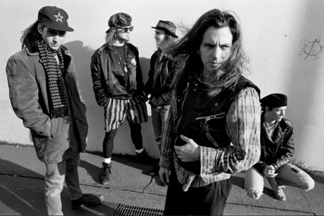 Five men with long hair and casual, grunge-style clothing stand and crouch against a plain wall outdoors, with one man in front looking directly at the camera and the others looking in different directions.