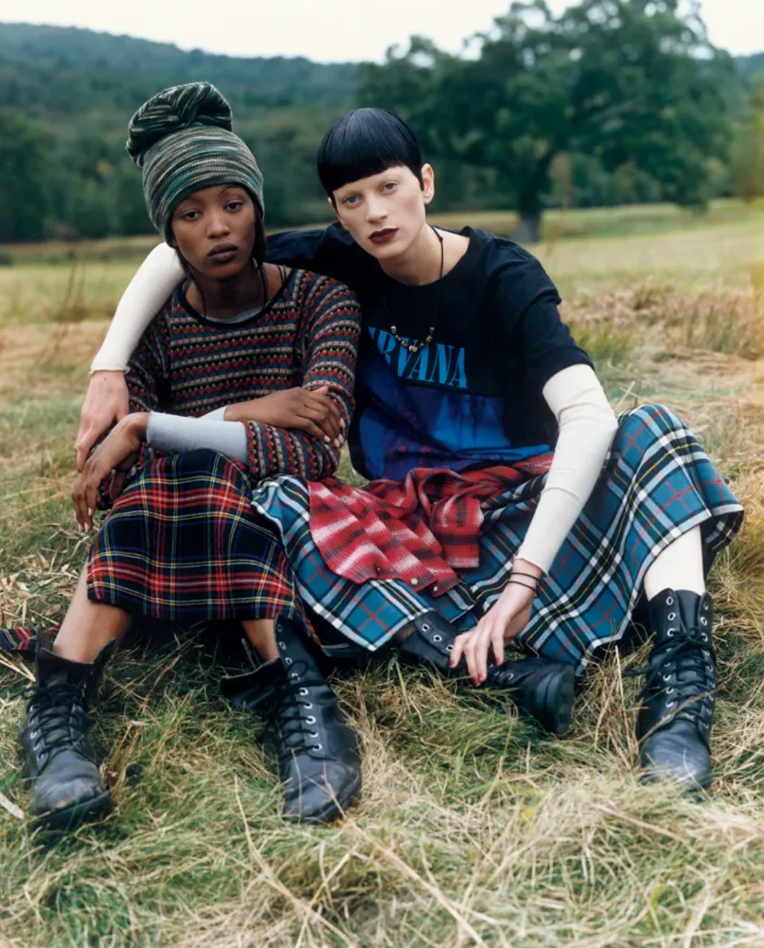 Two people sit on grass in a field, wearing patterned skirts, boots, and layered tops. One has a headwrap; the other has short dark hair and a Nirvana shirt. They pose with arms around each other, looking at the camera.