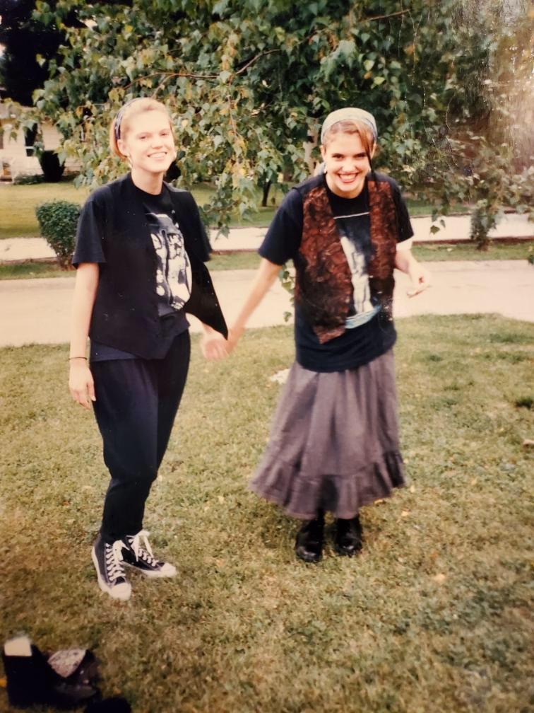 Two young women stand on grass, smiling and holding hands. One wears black pants, graphic tee, vest, and Converse sneakers; the other wears a long skirt, black boots, t-shirt, vest, and headband. Trees and a sidewalk are in the background.