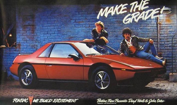 Two men with 1980s style hair and clothing pose with a red Pontiac sports car against a blue brick wall. The text says "MAKE THE GRADE" and "PONTIAC WE BUILD EXCITEMENT." A logo is visible in the corner.