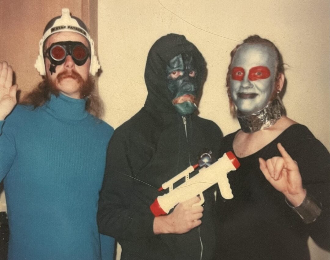 Three people in costumes pose indoors. One wears a helmet and blue shirt, another has a gorilla mask and holds a toy gun, and the third has silver face paint, a red mask, and a metallic collar.