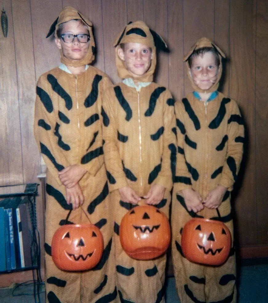 Three children stand indoors wearing matching tiger costumes and holding plastic pumpkin buckets, likely for Halloween. They are smiling and standing in front of a wood-paneled wall next to a small bookshelf.