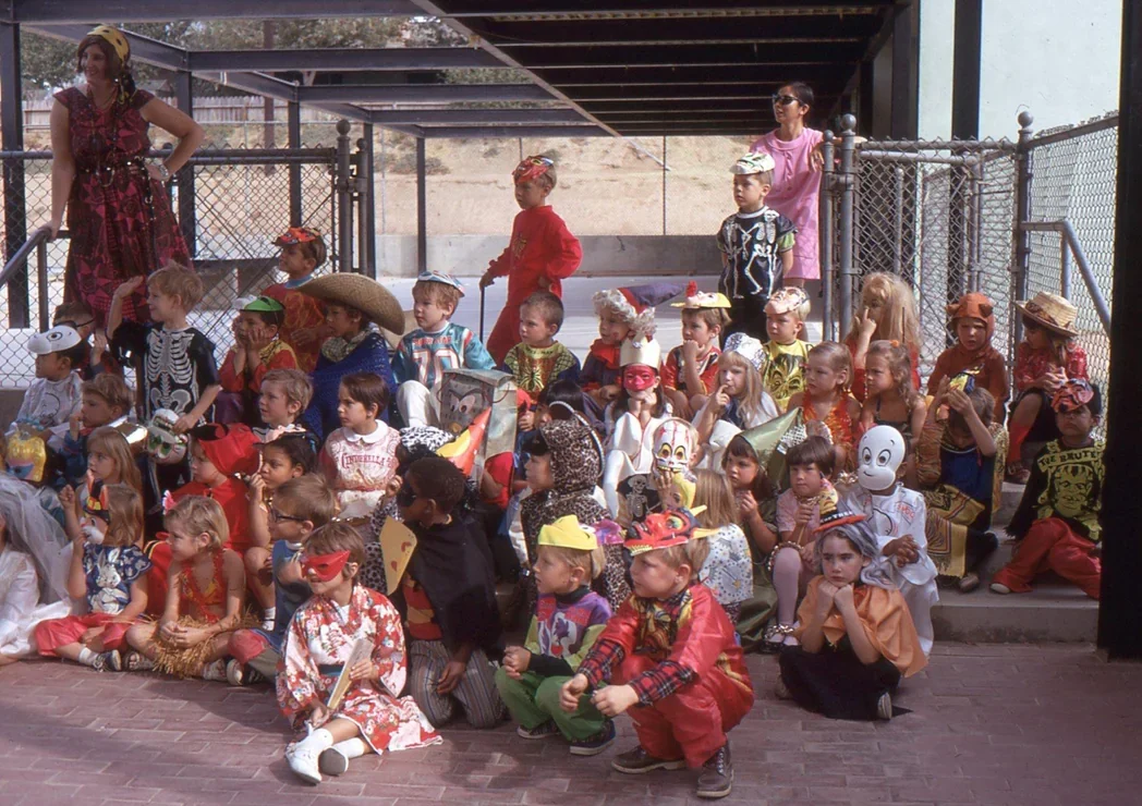 A large group of children in colorful costumes, including skeletons, clowns, and animals, sit and stand outside by a fence, posing for a group photo with two adults.