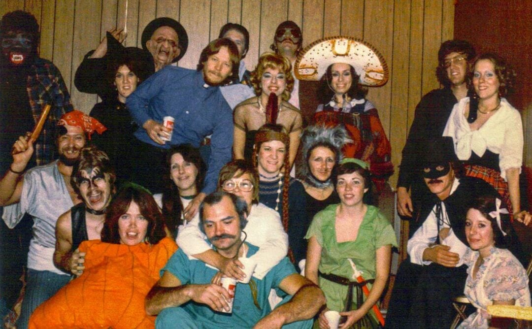 A group of adults in various colorful costumes pose together at a party, smiling in front of wood-paneled walls decorated with orange streamers.