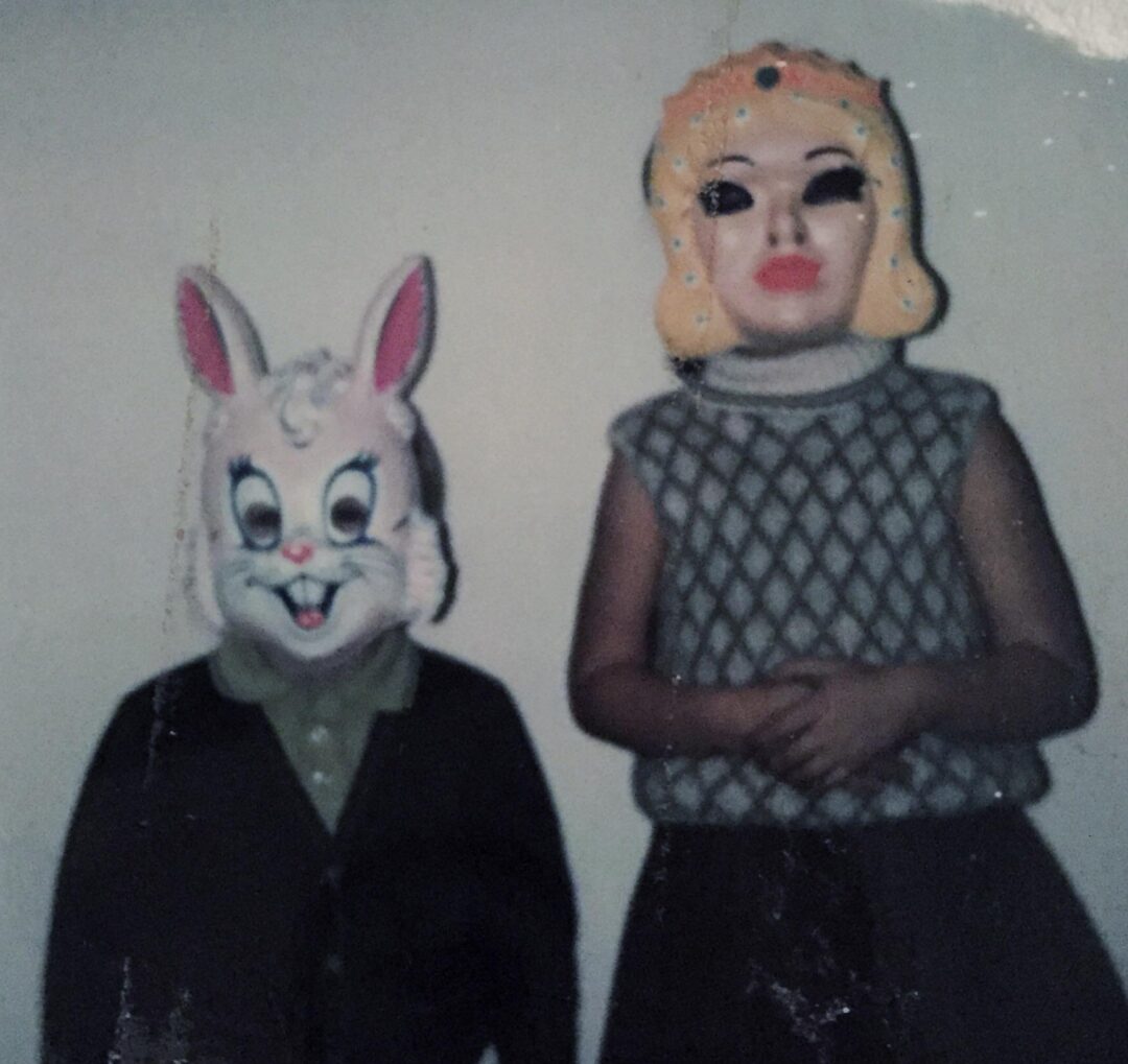 Two children stand side by side against a plain wall, wearing vintage Halloween masks—one a white rabbit with pink ears, the other a blonde woman. The photo is faded and partially damaged at the top.