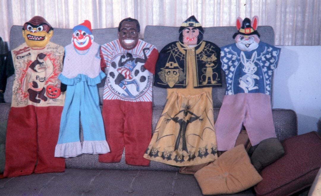 Five vintage Halloween costumes with colorful plastic masks and fabric outfits are displayed on a couch. The costumes include cartoon and clown characters, with pillows and cushions scattered on the side.