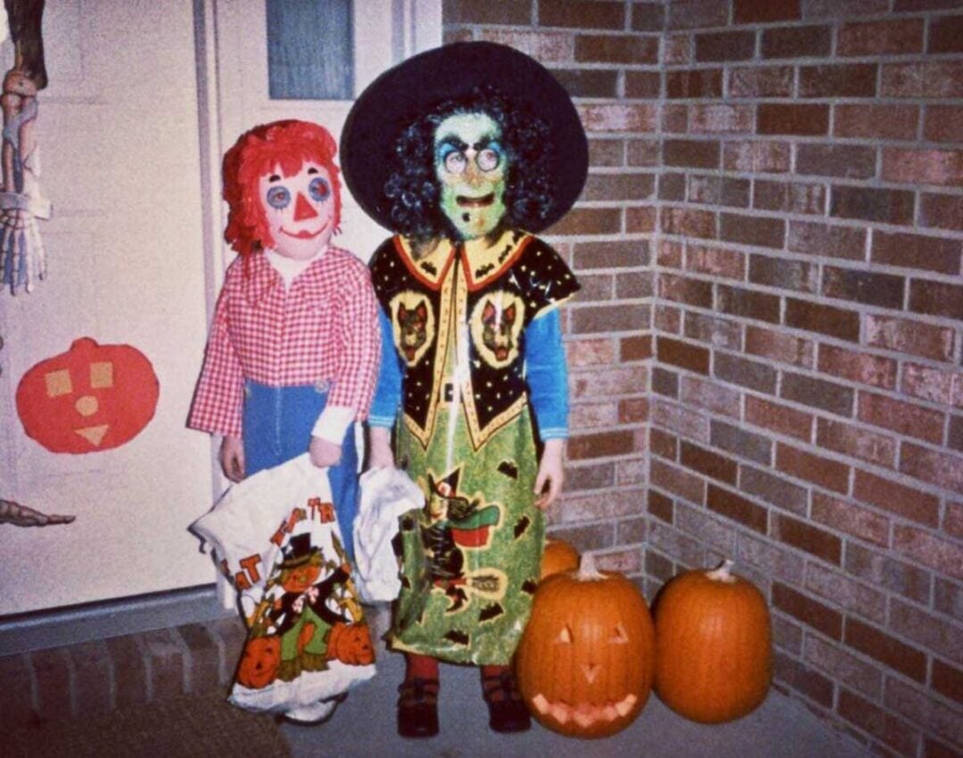Two children in Halloween costumes stand by a brick wall and pumpkins. One is dressed as a doll with red yarn hair and a checkered shirt, the other as a witch with a green face, glasses, and a black hat.