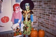 Two children in Halloween costumes stand by a brick wall and pumpkins. One is dressed as a doll with red yarn hair and a checkered shirt, the other as a witch with a green face, glasses, and a black hat.