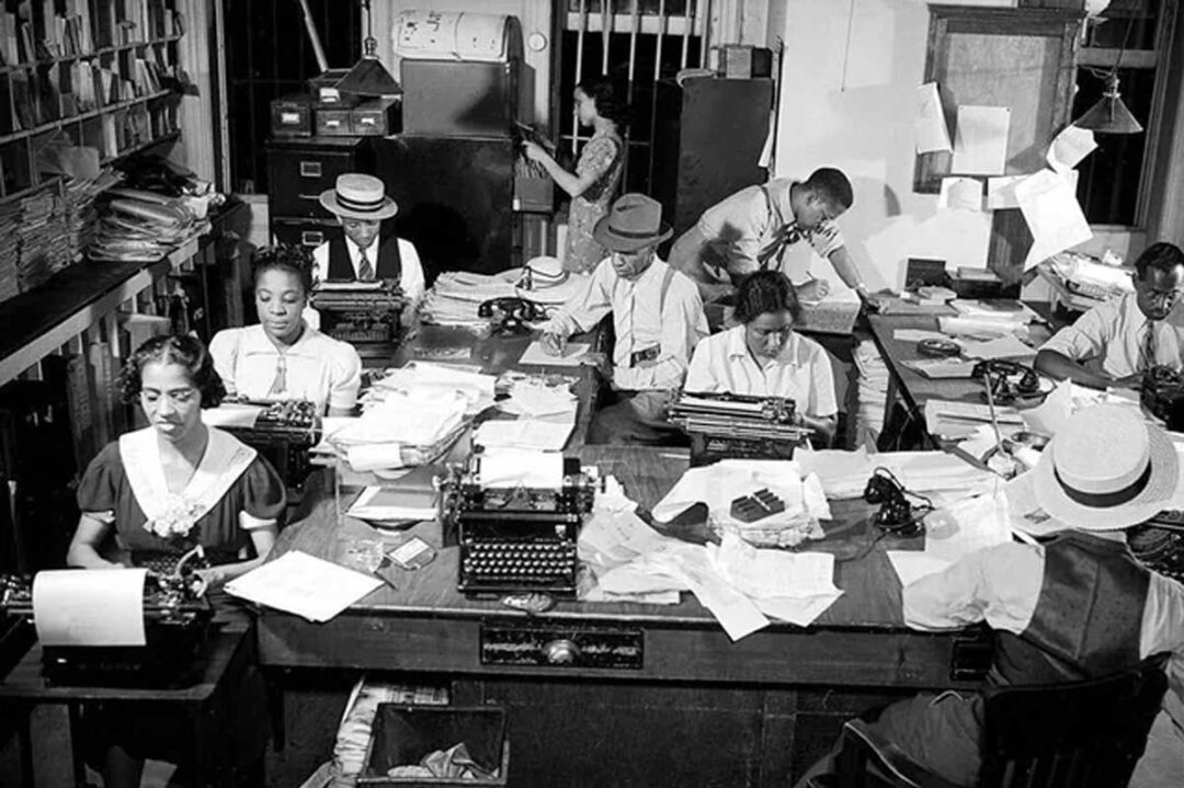 Black and white photo of several people typing at desks covered with papers and typewriters in a busy, crowded office. Shelves of files and papers hang on the walls in the background.