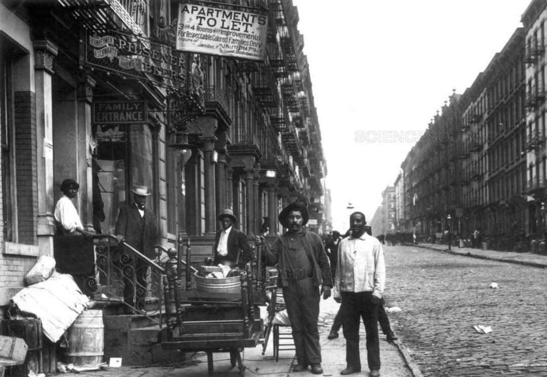 Black-and-white photo of men standing on a cobblestone street in an urban neighborhood, with apartment buildings, signs for apartments to let, and various items and barrels on the sidewalk.