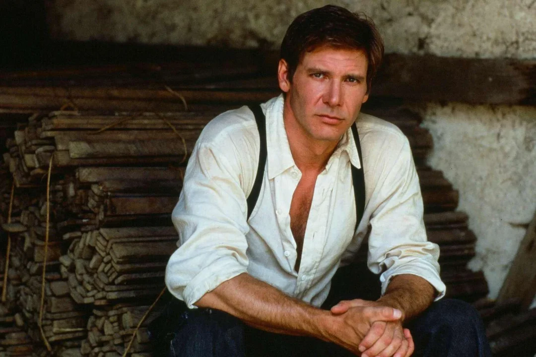 A man with short brown hair wearing a white button-up shirt with sleeves rolled up and black suspenders sits in front of stacked wooden planks, looking calmly at the camera.