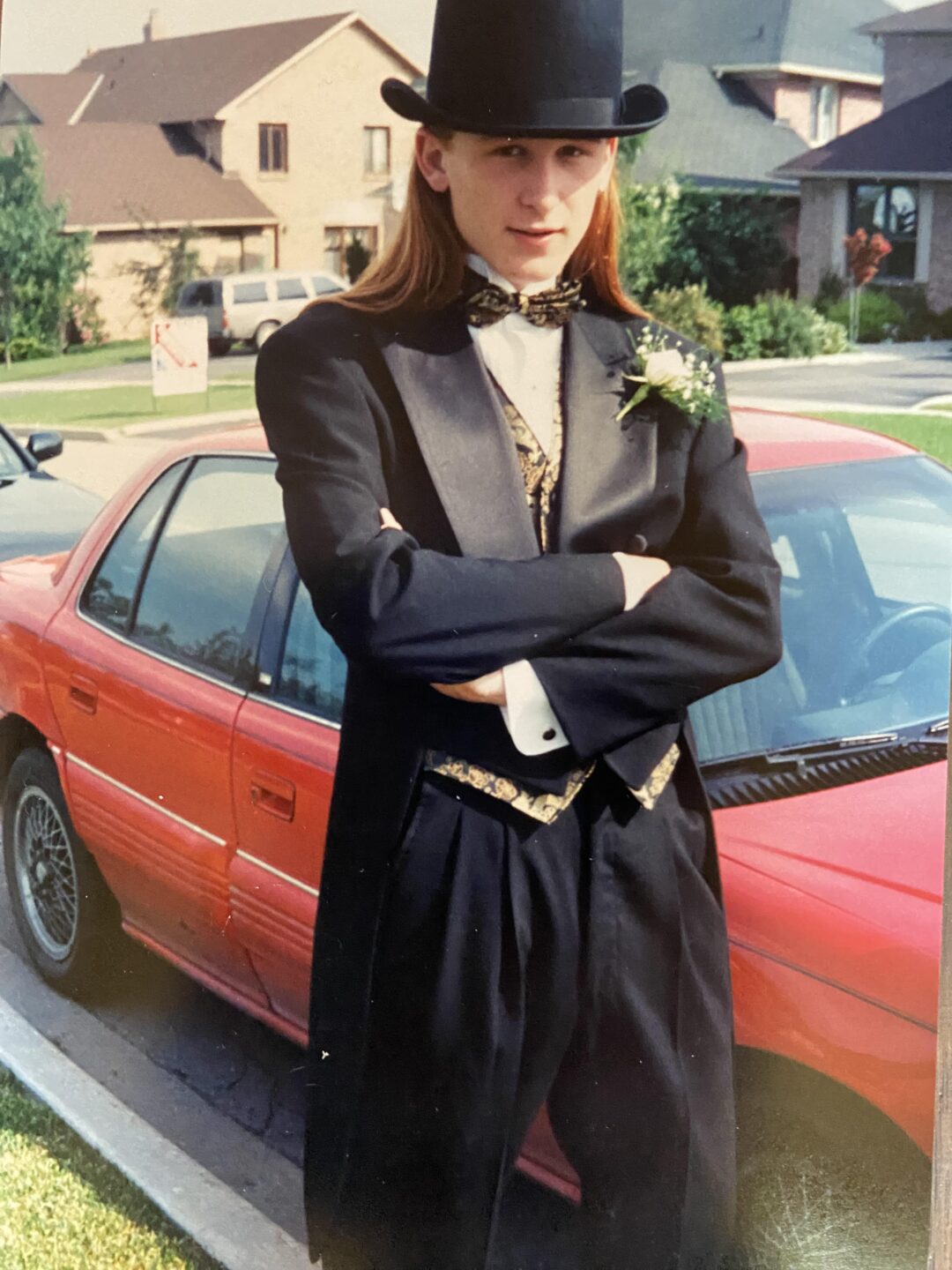 A young person with long hair, wearing a black tuxedo, gold and black vest, bow tie, and a top hat, stands with arms crossed in front of a red car on a suburban street.