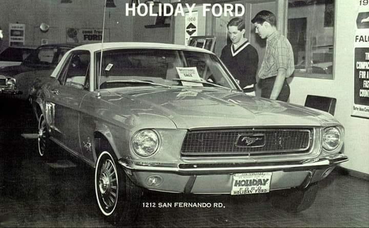 Black-and-white photo of two men looking at a classic Ford Mustang inside a dealership. A "Holiday Ford" sign is visible above, and the car has a "For Sale" sign and a "Holiday" license plate.