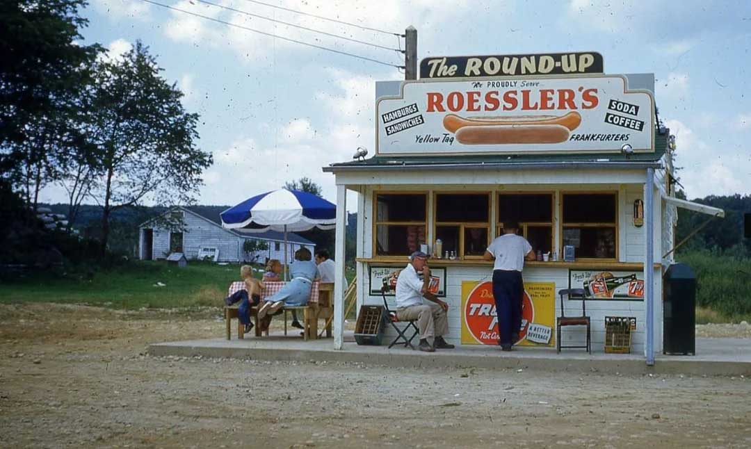 A small roadside hot dog stand named "The Round-Up" with a large Roessler’s hot dog sign. People sit at tables with umbrellas and a man stands at the service window. The scene is outdoors on a dirt lot with trees in the background.
