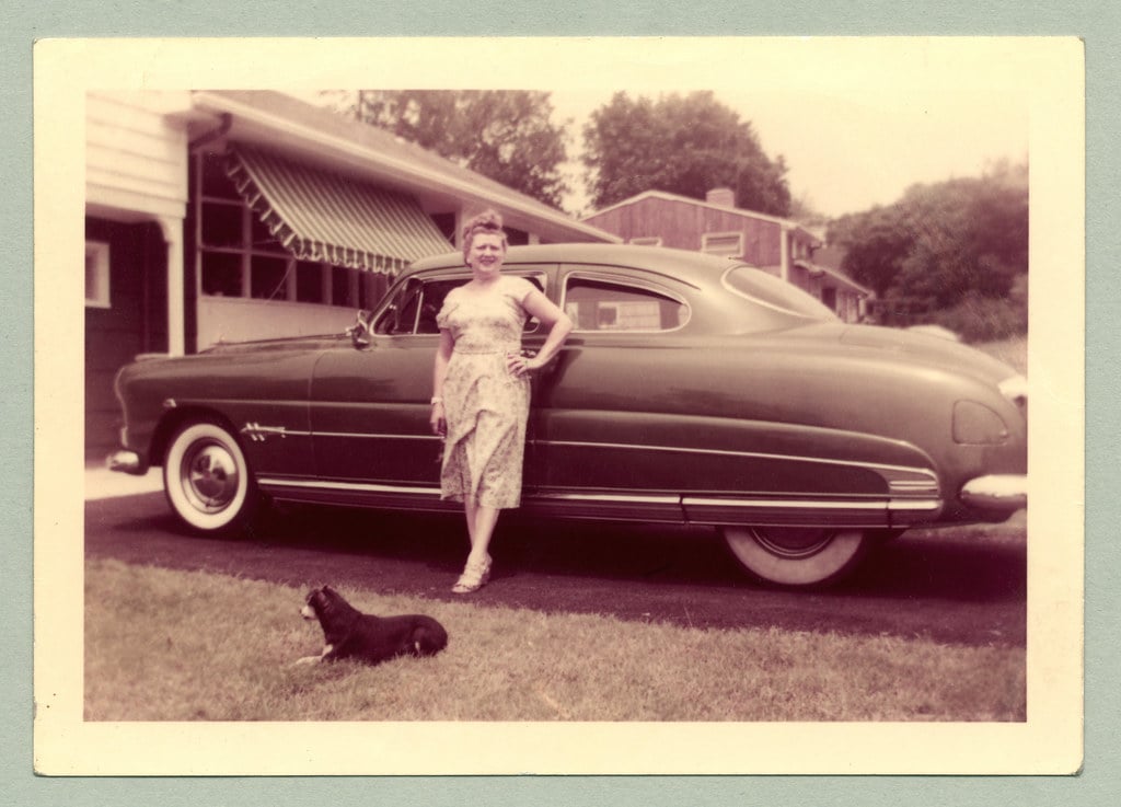 A woman in a 1950s-style dress stands by a vintage car parked in a driveway, with one hand on the car. A small dog sits on the grass in the foreground, and suburban houses are visible in the background.