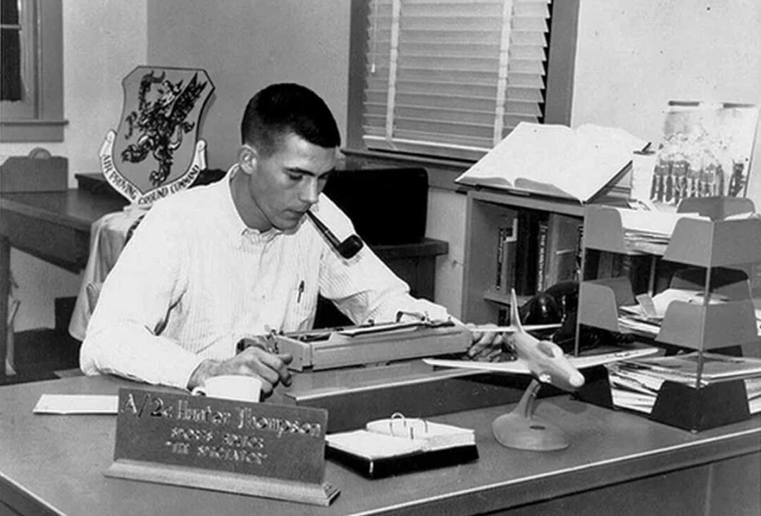 A young man sits at a desk typing on a typewriter in an office. The desk has a nameplate reading "A.V. Hunter Thompson," papers, a model airplane, and a binder. Shelves with books and a window are in the background.