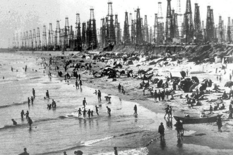 Crowded beach scene with many people swimming, playing, and relaxing along the shore. Numerous oil derricks and rigs line the background, creating a stark contrast with the seaside activity. Black-and-white, historical photo.
