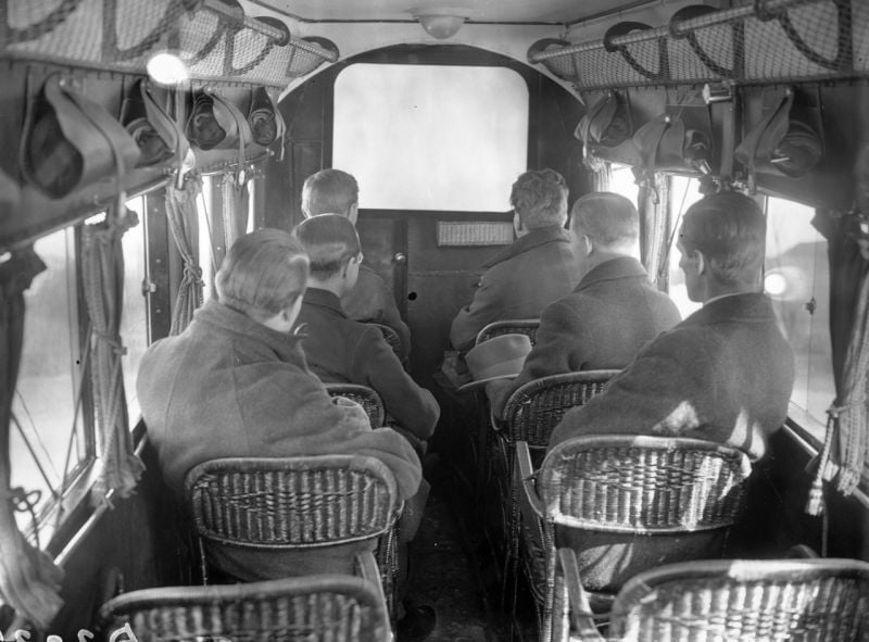 Six men with short hair, wearing coats, sit in wicker seats inside an old-fashioned vehicle or aircraft, facing forward toward a partition with windows. Overhead racks hold bags, and daylight comes through side windows.