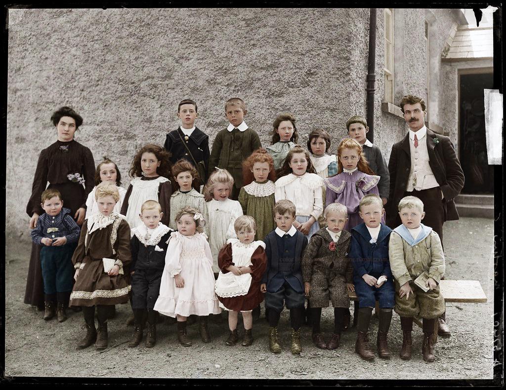 A vintage photo of a group of children and two adults in old-fashioned clothing, posed outside against a stone wall. The children are arranged in three rows, with some standing and some sitting.