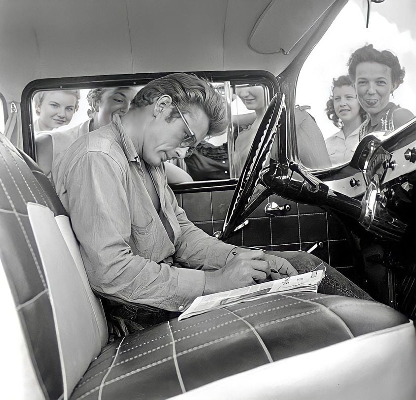 A man with glasses sits in a car, signing autographs on a notepad, while a group of smiling young women look at him through the open window. The scene appears to be from the mid-20th century.