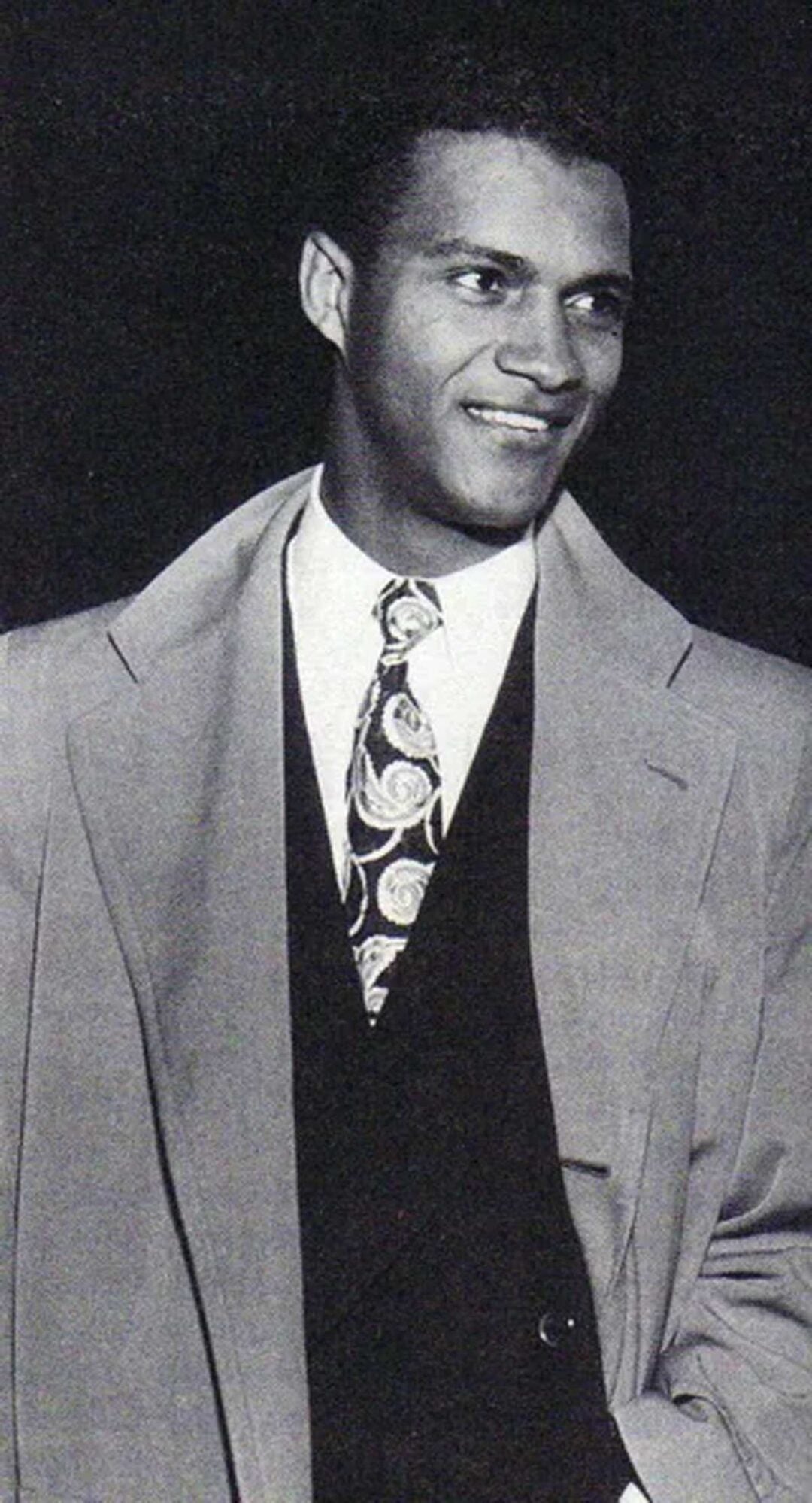 A man dressed in a suit, patterned tie, and light-colored overcoat smiles while looking to the side. The image is black and white.