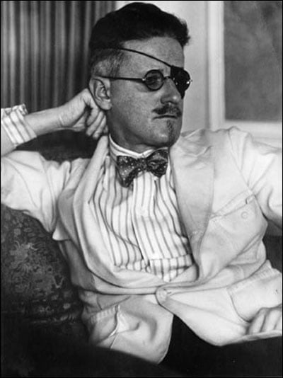 A man wearing an eye patch, round glasses, and a light-colored suit with a striped shirt and bow tie sits with one arm resting on a chair, gazing thoughtfully to the side.