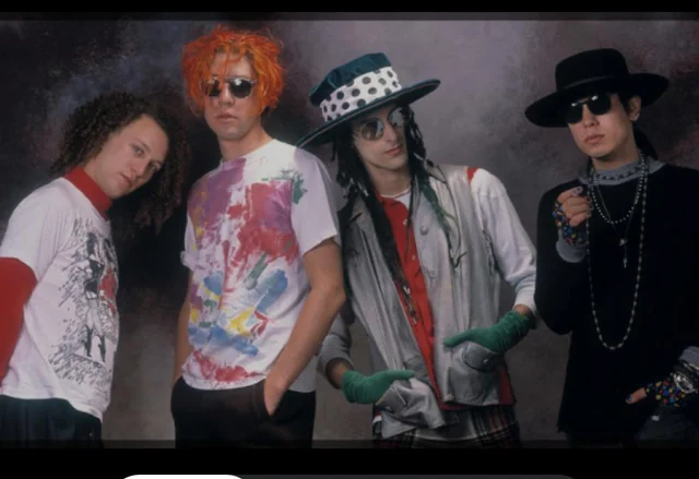 Four men pose together in colorful, eclectic outfits; one has curly hair, another bright orange hair and sunglasses, the third wears a large polka-dot hat and green gloves, and the fourth wears all black with dark sunglasses and a wide-brimmed hat.