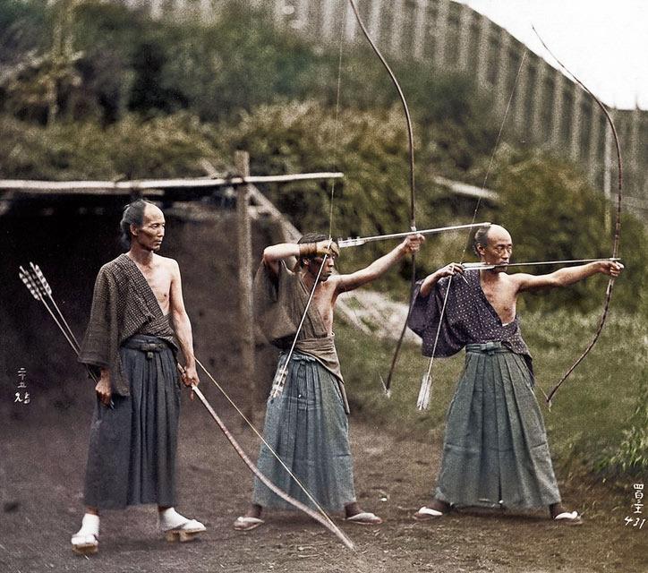 Three men in traditional Japanese clothing stand outdoors; two are drawing longbows while the third holds arrows, watching. All are barefoot and wear loose pants, with greenery and a wooden structure in the background.