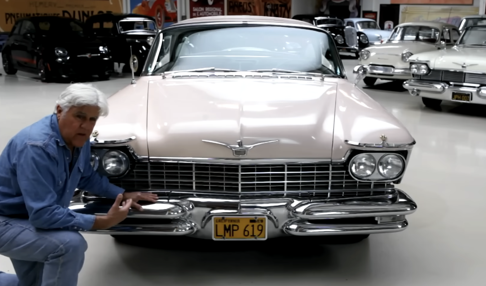 A man in a blue shirt crouches next to a vintage beige car with chrome detailing and a yellow license plate reading “LMP 619” inside a classic car showroom.