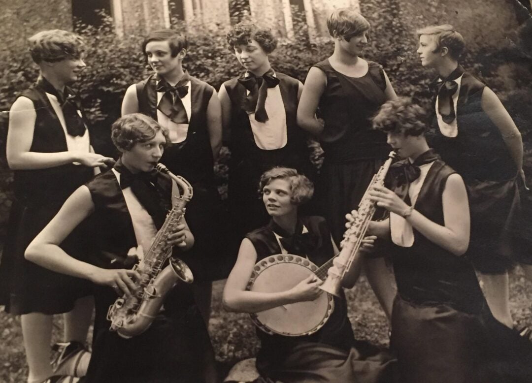 Eight women in 1920s-style dresses pose outdoors; two play saxophones, one plays a banjo, and others smile and look at each other, suggesting they are members of a jazz band.